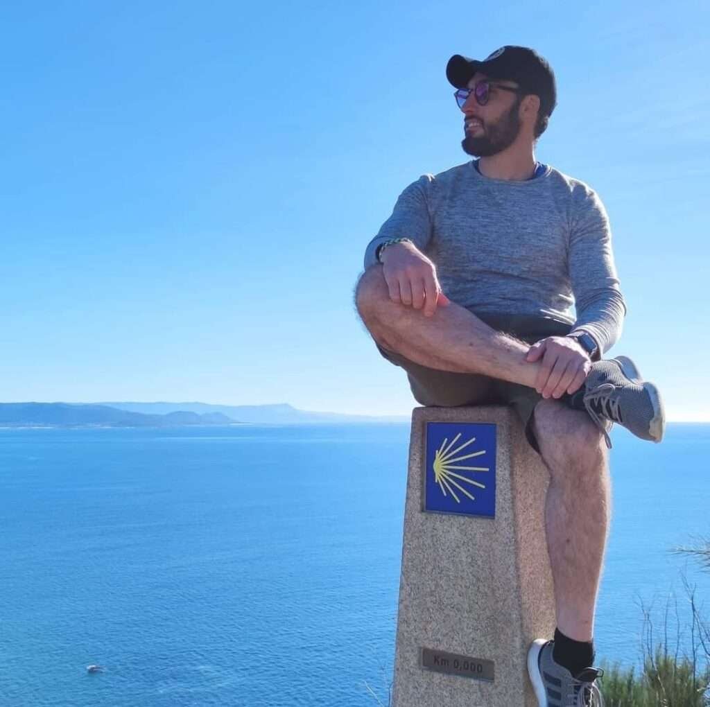 Giovanni Pagoria seduto sulla colonna del km 0 dopo il viaggio di Santiago, con vista sul mare.