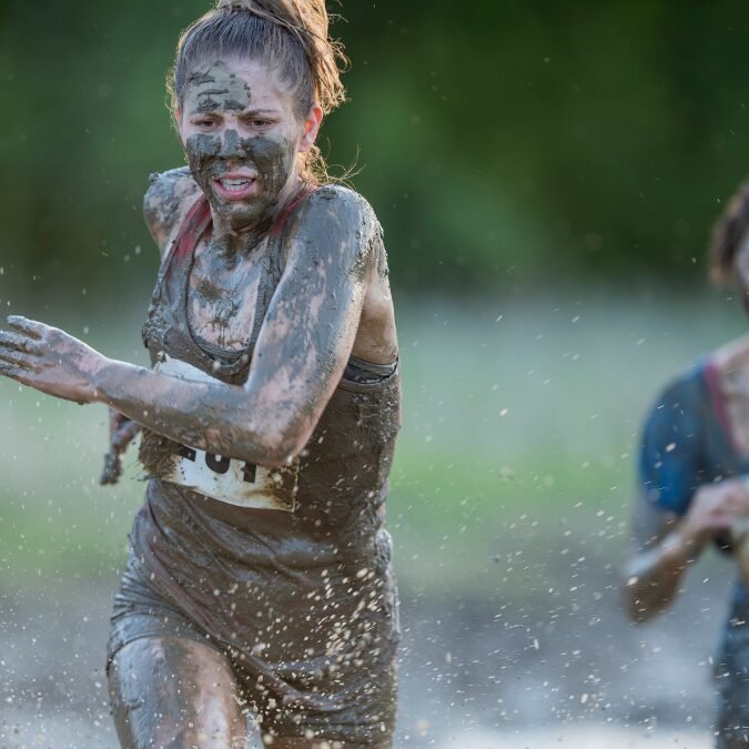 Una giovane donna corre nel fango durante una gara ad ostacoli. Il suo viso e il corpo sono completamente sporchi di fango, ma l’espressione determinata e concentrata rivela come essere costanti nella vita per raggiungere gli obiettivi. 